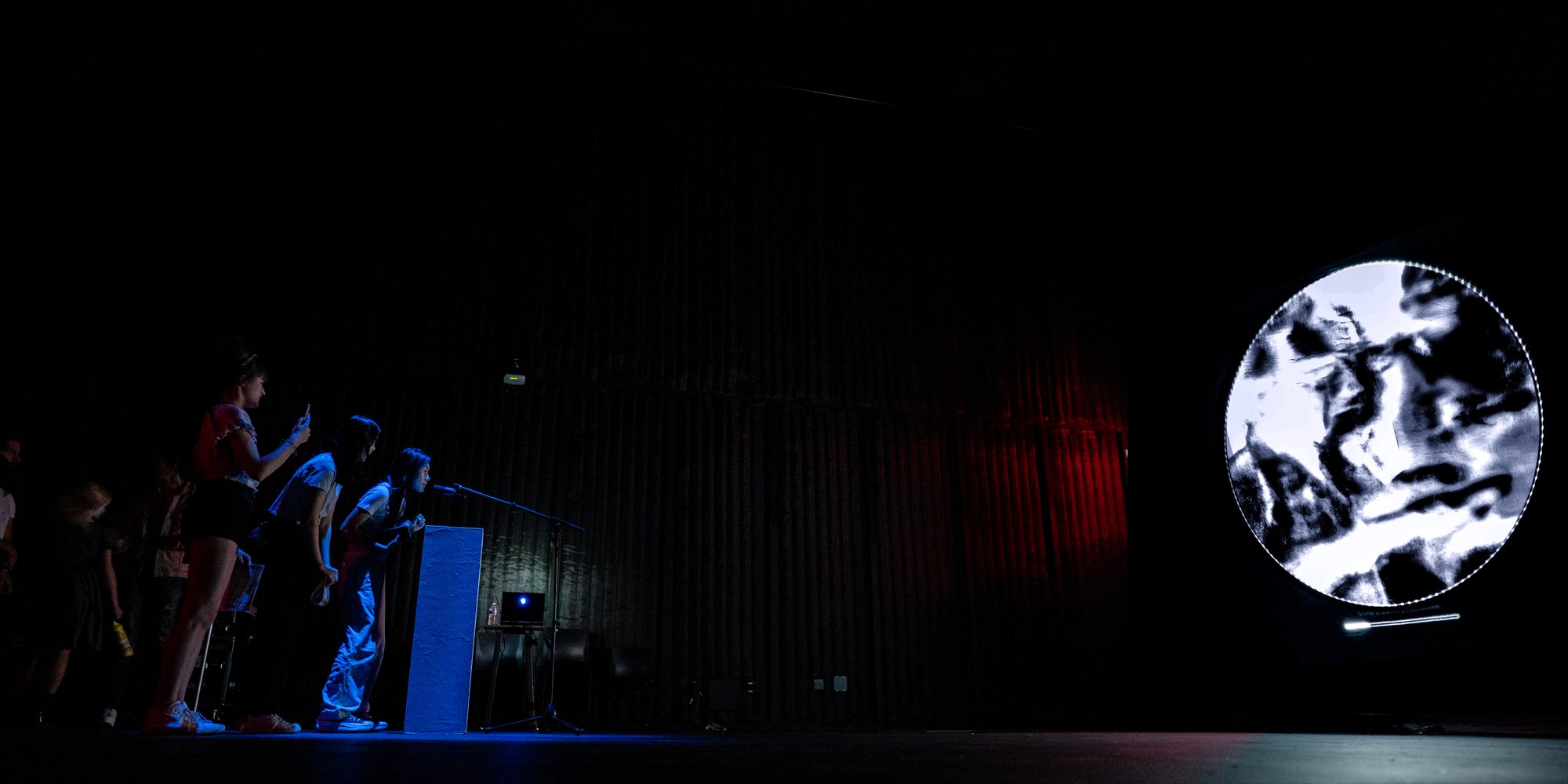 The installation in a dark room: a round screen showing a generated memory, a red podium with the microphone, visitors watching